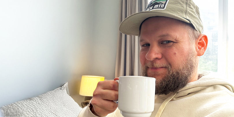 A bearded person in a beige hoodie and cap is sitting on a bed, smiling and holding a white mug. There is a lit lamp and a light gray wall in the background. Natural light comes in through a window.