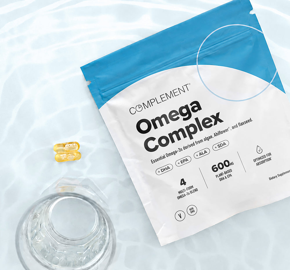 A packet labeled Omega Complex with supplement details lies on a light surface near two yellow capsules and a glass of water.