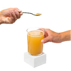 Two hands appear against a plain background. One hand holds a clear glass of orange juice on a small white pedestal, while the other hand holds a spoon with powder, poised above the glass.