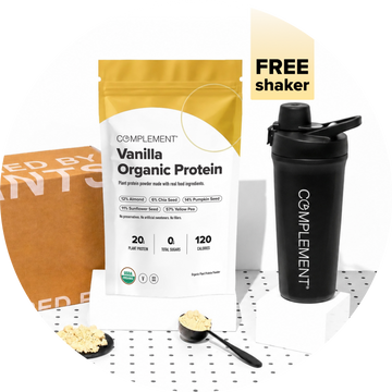 A bag of Complement Vanilla Organic Protein powder, a black shaker bottle with Complement branding, a cardboard box, and two spoons with protein powder are arranged on a white polka-dot surface. A FREE shaker label is shown.