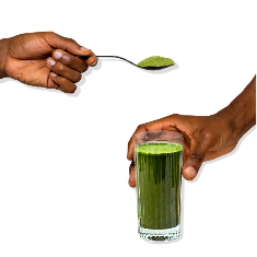 A hand holds a glass filled with green liquid, while another hand holds a spoon with green powder above it, both set against a plain white background.