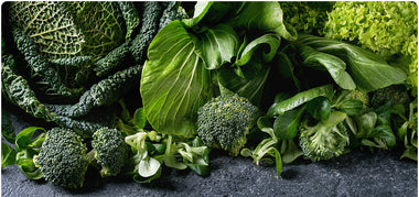 A variety of fresh green vegetables, including broccoli, kale, bok choy, and lettuce, arranged closely together on a dark stone surface.
