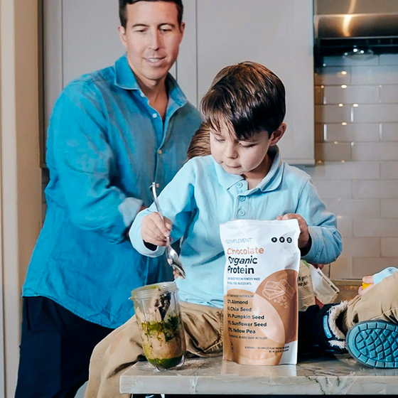 A young boy scoops a green smoothie from a blender jar on a kitchen counter, with a packet of chocolate organic protein powder beside him. An adult stands nearby, both wearing blue shirts.