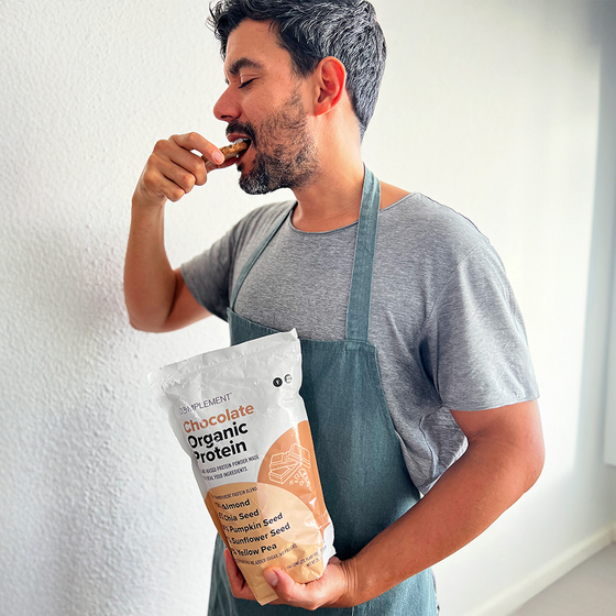 A man wearing a gray t-shirt and apron holds a bag of chocolate organic protein powder while tasting some with his fingers, standing against a white wall in a bright room.