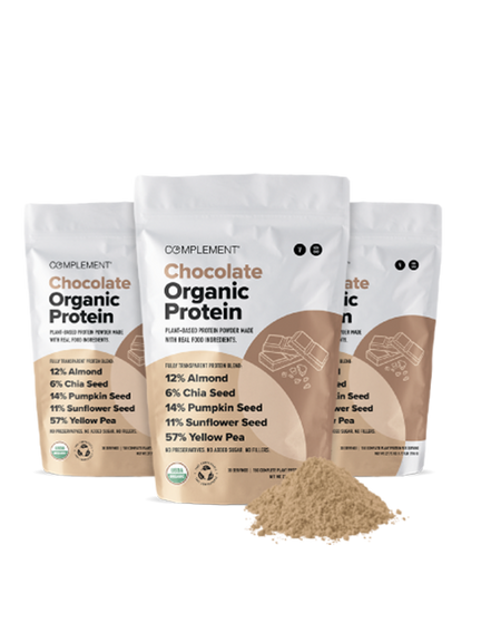 Three bags of Complement Chocolate Organic Protein powder with plant-based ingredients like almond, chia seed, and pumpkin seed. A small pile of the protein powder is in front of the bags.
