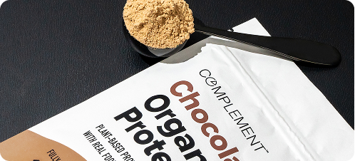 A black spoon with protein powder rests on a dark surface next to an open white packet labeled “Complement Chocolate Organic Protein.”.