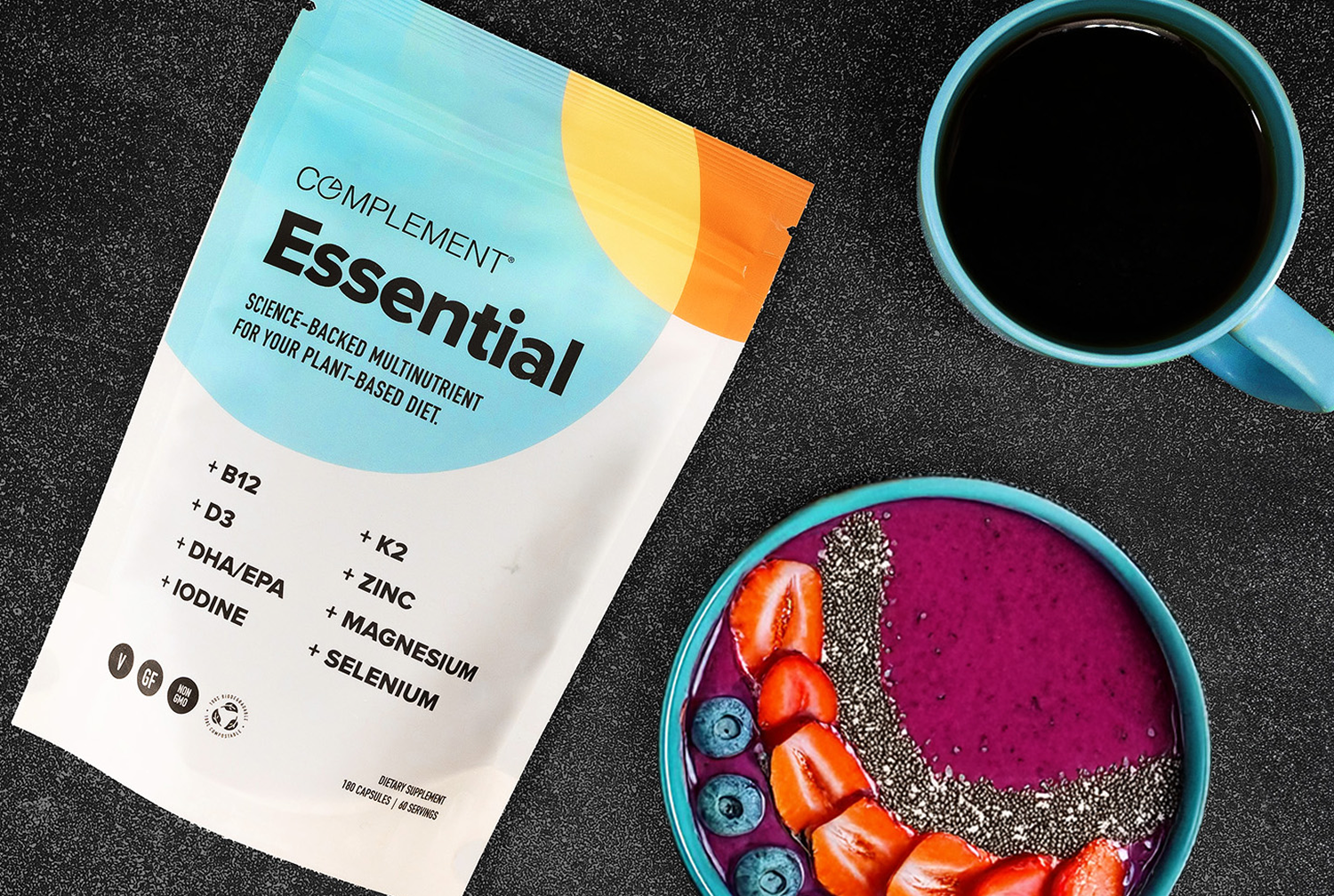A bag of Complement Essential multinutrient powder sits on a black surface next to a blue mug filled with coffee and a smoothie bowl topped with strawberries, blueberries, chia seeds, and granola.