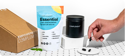 A hand picks up black capsules from a white dish next to a pouch labeled Essential and a black container, with a brown box labeled Empowered by Complement in the background.