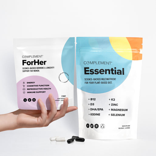 Two supplement pouches labeled Complement ForHer and Complement Essential stand upright on a white surface. A hand holds the ForHer pouch. Several black and white capsules and pills lie in front.