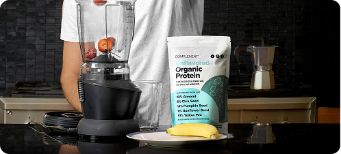 A person stands in a kitchen behind a blender with fruit inside, a glass, a white package labeled Organic Protein, and a plate with a peeled banana in the foreground.
