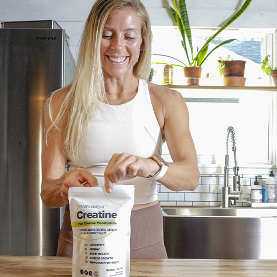 A woman in a kitchen opens a bag of creatine supplement. Shes wearing a white tank top and light pink pants. In the background are kitchen shelves, a plant, and a stainless steel fridge. Shes smiling and has a smartwatch on her wrist.