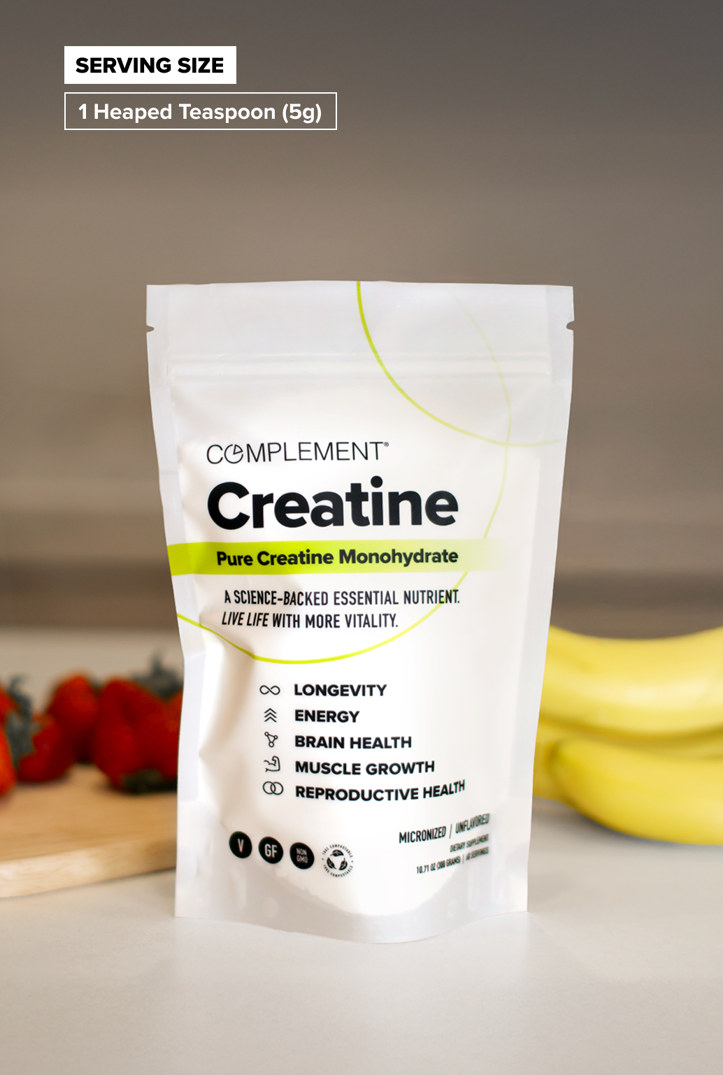 A white bag labeled Complement Creatine stands on a counter with strawberries and bananas in the background. The packaging highlights benefits like longevity, energy, brain health, muscle growth, and reproductive health.