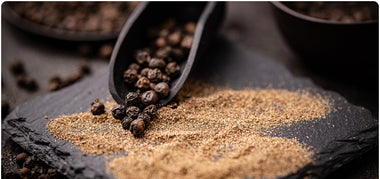 Whole black peppercorns spill from a scoop onto a dark stone surface, next to scattered piles of ground black pepper.