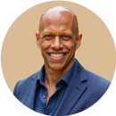 A smiling bald man wearing a dark blue blazer and blue shirt, posed against a plain beige background.