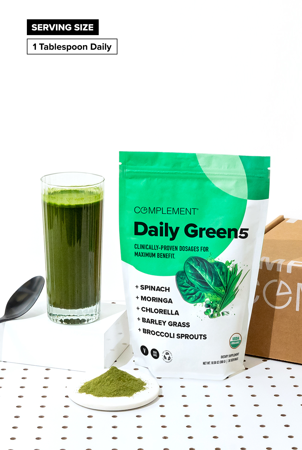 A bag of Complement Daily Greens powder stands next to a glass of green drink, a spoon, and a small heap of the powder on a white surface. A label reads Serving Size: 1 Tablespoon Daily.