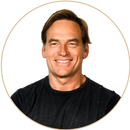 Smiling adult man with light brown hair wearing a black t-shirt, photographed against a light gray background within a circular frame.