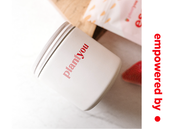 A white container with planti you in red text on its side sits on a light surface. Nearby, theres partially visible packaging and a strawberry. A wooden board peeks out in the background.