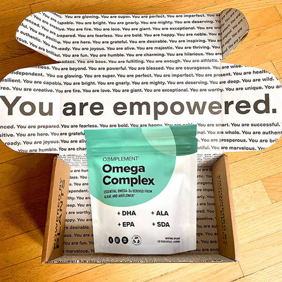 A package of Complement Omega Complex supplement sits open on a wooden floor. The inner lid of the box displays positive affirmations, with the prominent message You are empowered in bold.