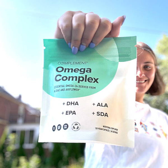 A person smiles while holding up a Complement Omega Complex supplement pouch. The pouch is green and white, and includes text about omega-3 derived from algae and ahiflower. The background features a sunny outdoor scene.