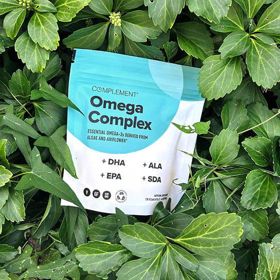 A packaging of Complement Omega Complex lies on green leaves. The bag is teal and white, displaying text about essential Omega-3 derived from algae and Ahiflower, along with +DHA, +EPA, +ALA, and +SDA.