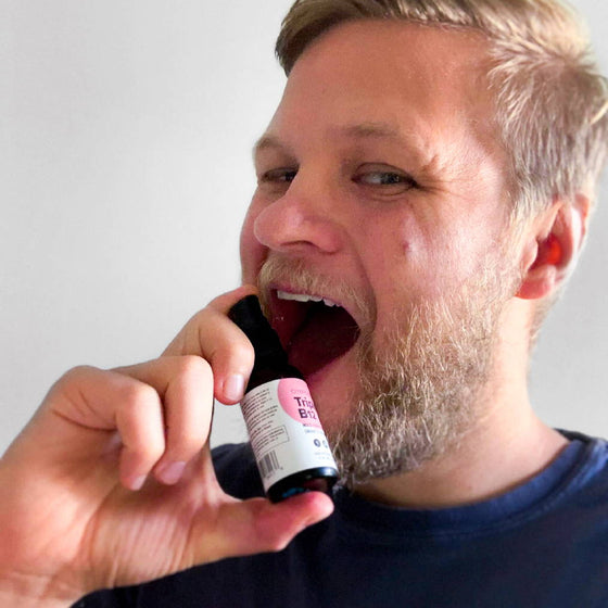 A person with a beard is playfully pretending to drink from a small bottle labeled Cider Extract. They are wearing a dark shirt and have a light, neutral background. The persons expression is cheerful.