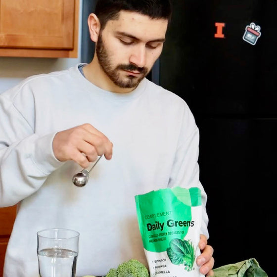 Man in a white sweatshirt scoops green powder from a Daily Greens bag into a glass of water. A broccoli is on the counter, and a fridge with a magnet is in the background.