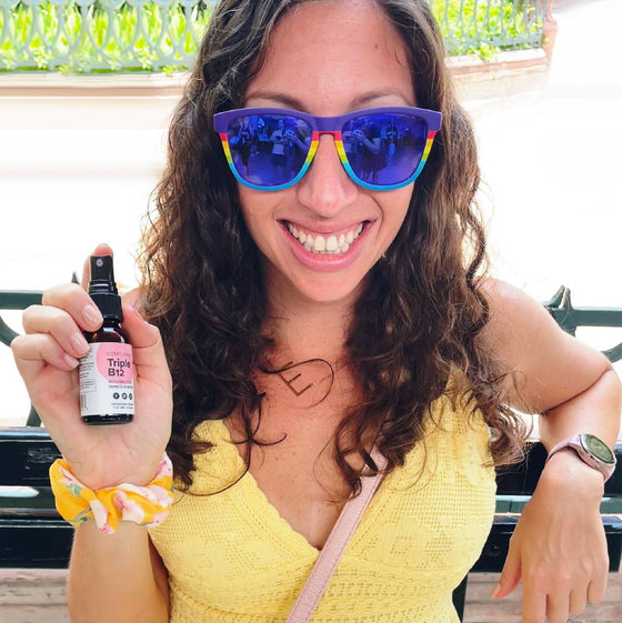 A person with long curly hair and wearing colorful sunglasses smiles at the camera. They are holding a small bottle labeled Triple B12. Wearing a yellow sleeveless top and a scrunchie on their wrist, they sit outdoors on a sunny day.