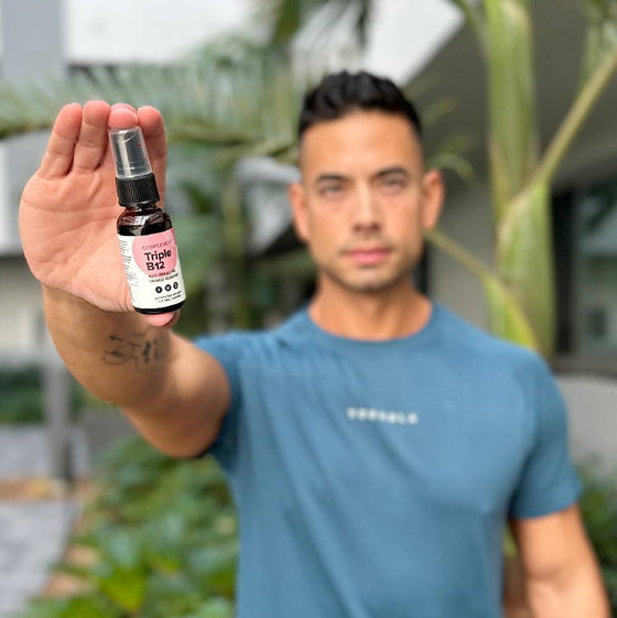 A person in a blue shirt holds a spray bottle labeled Triple B12 towards the camera, standing in an outdoor setting with blurred greenery and a building in the background.