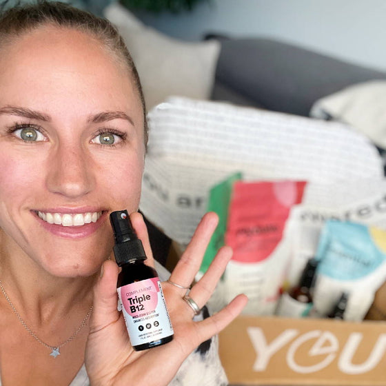 A person smiling and holding a bottle labeled Triple B12 spray. In the background, a box filled with various health products is visible. The letters YOU are partially seen on the box.