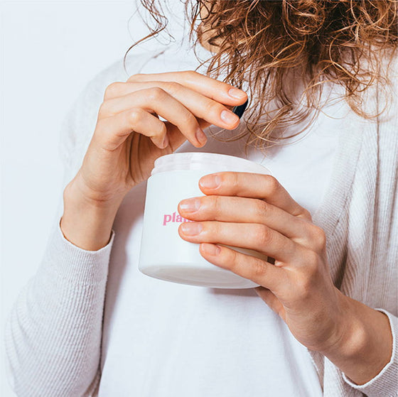 A person with curly hair is holding and opening a white jar of hair product. They are wearing a white top, and the background is plain. The brand name on the jar is partially visible.