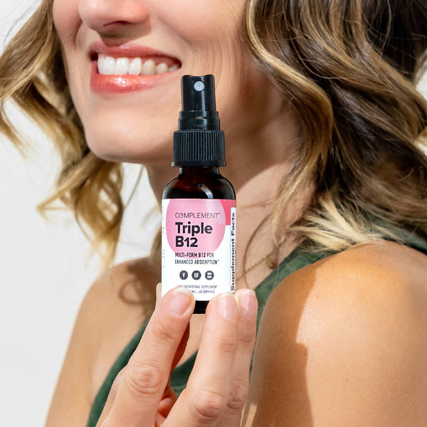 A person smiling and holding a small spray bottle labeled Complement Triple B12 with a pink and white design. The focus is on the product, and the person is wearing a green top.