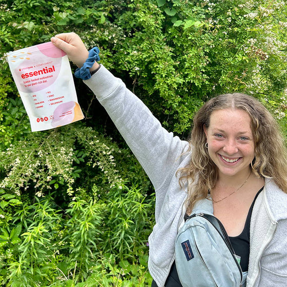 A smiling person stands outside in front of lush greenery, holding up a pink and white package labeled essential. Theyre wearing a light gray hoodie and carrying a light blue bag.