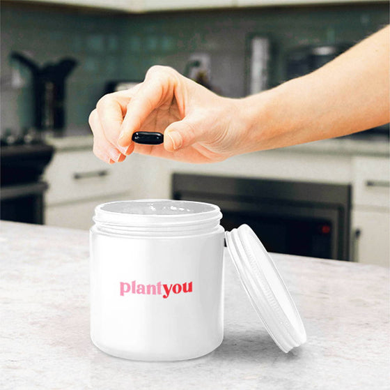 A hand holds a capsule above an open white jar labeled plantyou on a kitchen counter. The kitchen has a stove and cabinets in the background.