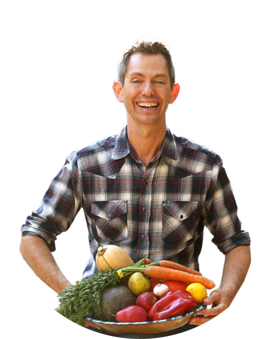 A person in a plaid shirt is smiling while holding a bowl filled with a variety of fresh vegetables, including carrots, bell peppers, a potato, and a squash, against a plain white background.