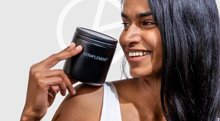 A person with long dark hair smiles while holding a black jar labeled COMPLEMENT against a light background.