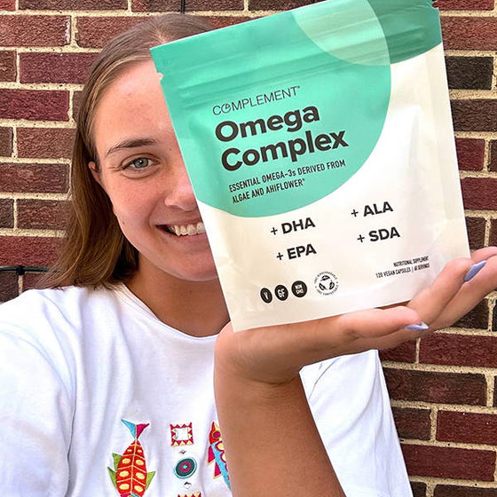 A person smiling and holding a package of Complement Omega Complex, which contains essential omega-3s derived from algae and ahiflower. The package lists DHA, EPA, ALA, and SDA as ingredients. A brick wall is in the background.