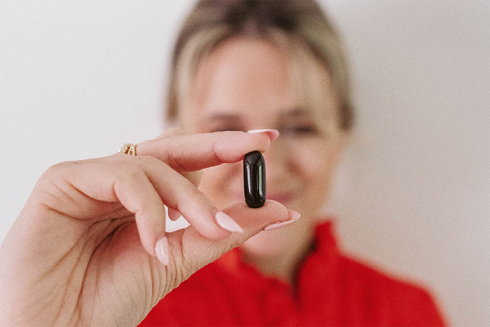 A person in a red shirt holds a small, dark capsule close to the camera, with fingers gently pinching it. The persons face is slightly blurred in the background.