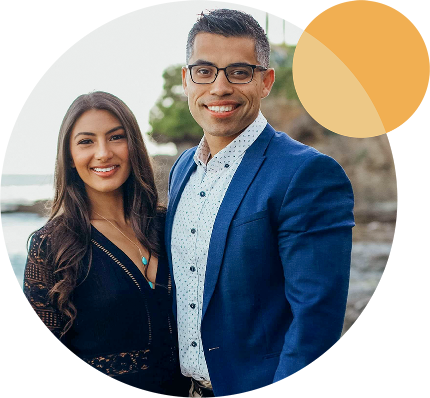 A smiling couple poses outdoors near a rocky shoreline. The man wears glasses, a blue blazer, and a patterned shirt, while the woman is in a black blouse with wavy hair. A circular orange graphic decorates the top right corner.
