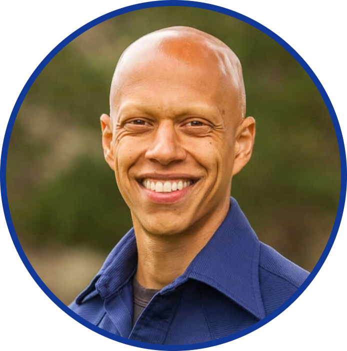 A smiling person with a shaved head wearing a blue collared shirt, set against a blurred outdoor background.