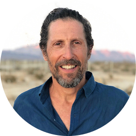 A man with short curly brown hair and a beard, wearing a blue shirt, smiles outdoors with a desert landscape and mountains in the blurred background.