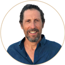 Smiling middle-aged man with short curly hair, beard, and mustache, wearing a blue button-up shirt, seen against a plain light background within a circular frame.