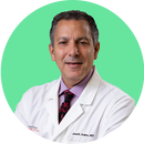A middle-aged man wearing a white lab coat, dress shirt, and patterned tie, smiling in front of a solid mint green background. The name Joel K. Kahn, MD is visible on his coat.