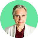 A senior woman with short gray hair wears a white blazer over a burgundy shirt, smiling softly in front of a solid green background.