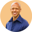 A smiling bald man wearing a blue collared shirt stands in front of a solid light orange background.