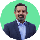 A man with short dark hair, a beard, and mustache, wearing a blue suit jacket and light blue shirt, smiles in front of a plain light green circular background.