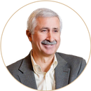 A middle-aged man with short gray hair and a mustache, wearing a gray suit jacket over a light-colored shirt, smiling against a plain light background.