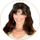 Smiling woman with long, wavy brown hair wearing a beige top, hoop earrings, and a circular necklace, posed against a light gray background with a circular border.