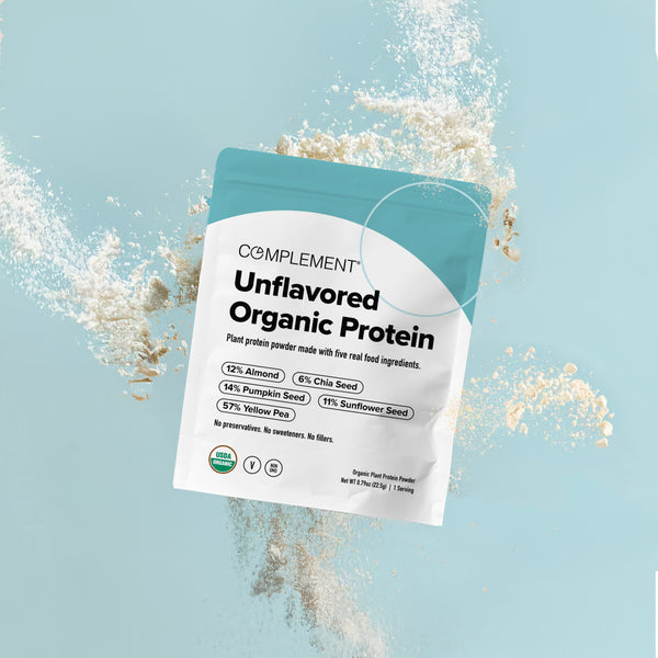 A bag of Complement Unflavored Organic Protein powder sits upright, surrounded by scattered powder. The packaging highlights its plant-based, organic ingredients and contains info about almond, pumpkin, chia, sunflower, and yellow pea content.