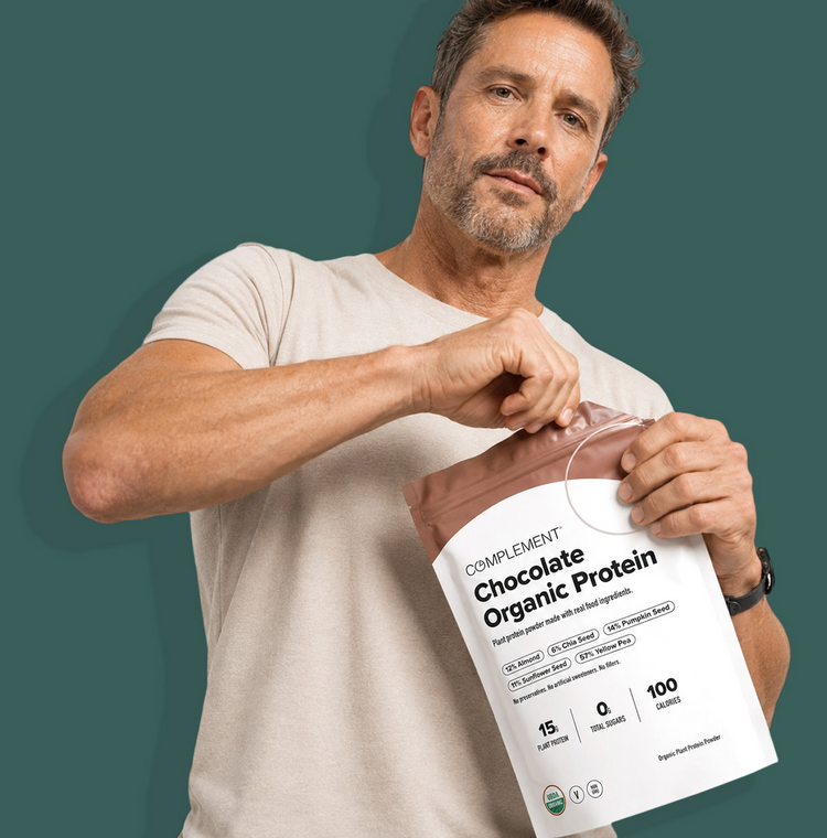 A man in a beige t-shirt opens a bag labeled Complement Chocolate Organic Protein against a solid green background. The packaging highlights 15g protein, 0g sugar, and 100 calories per serving.
