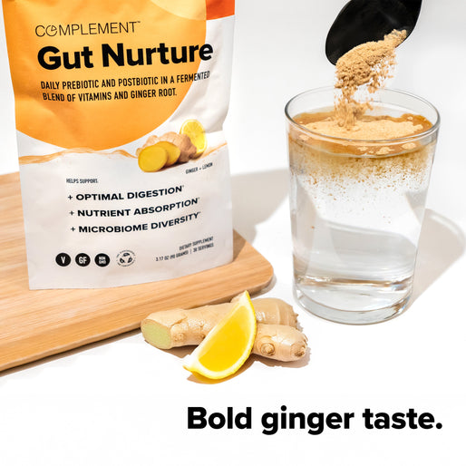 A packet of Complement Gut Nurture powder stands next to a glass of water as powder is poured in. Fresh ginger and a lemon wedge rest on a wooden board. Text reads: Bold ginger taste.